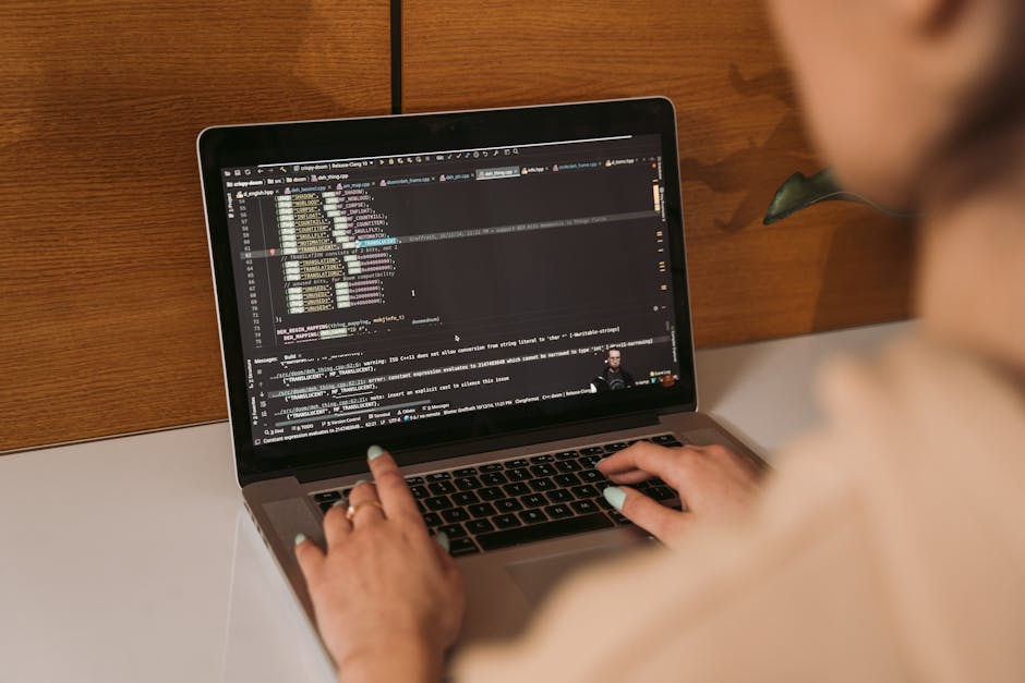 A person working on a laptop placed on a white desk against a wooden background, with the screen displaying lines of code in a dark-themed text editor. The individual's hands are visible, with one han